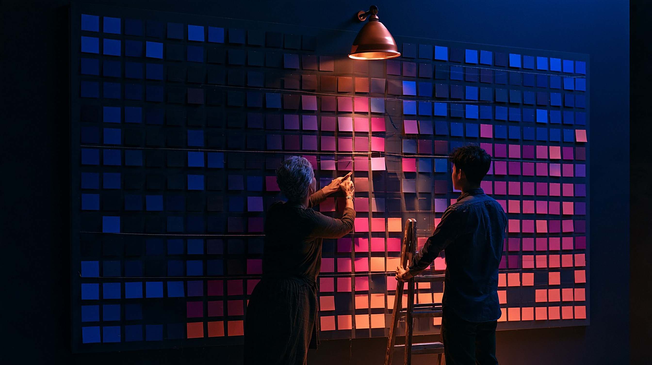 Two people arrange a large wall grid of coloured cards under a copper lamp.