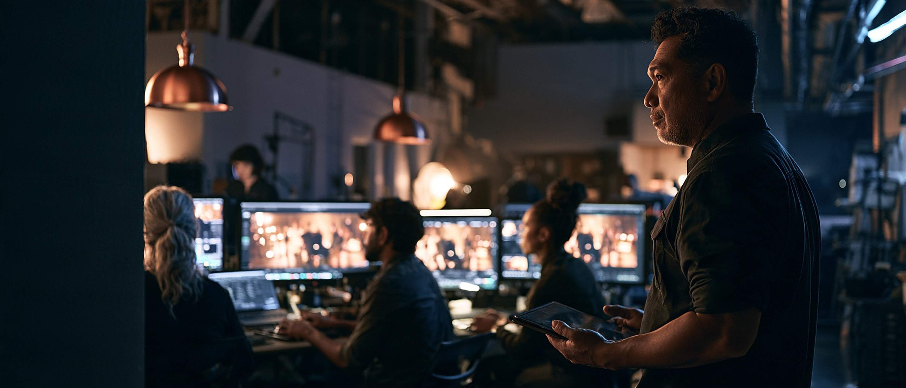 A post-production supervisor holds a tablet and looks across a studio where editors work at glowing screens.