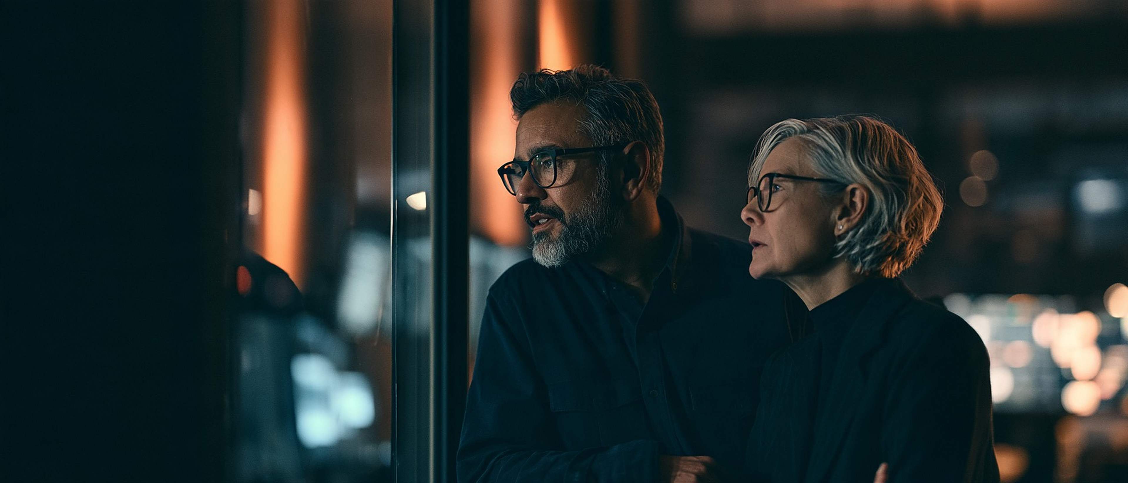 Two senior producers talk beside a glass wall overlooking an editing suite.