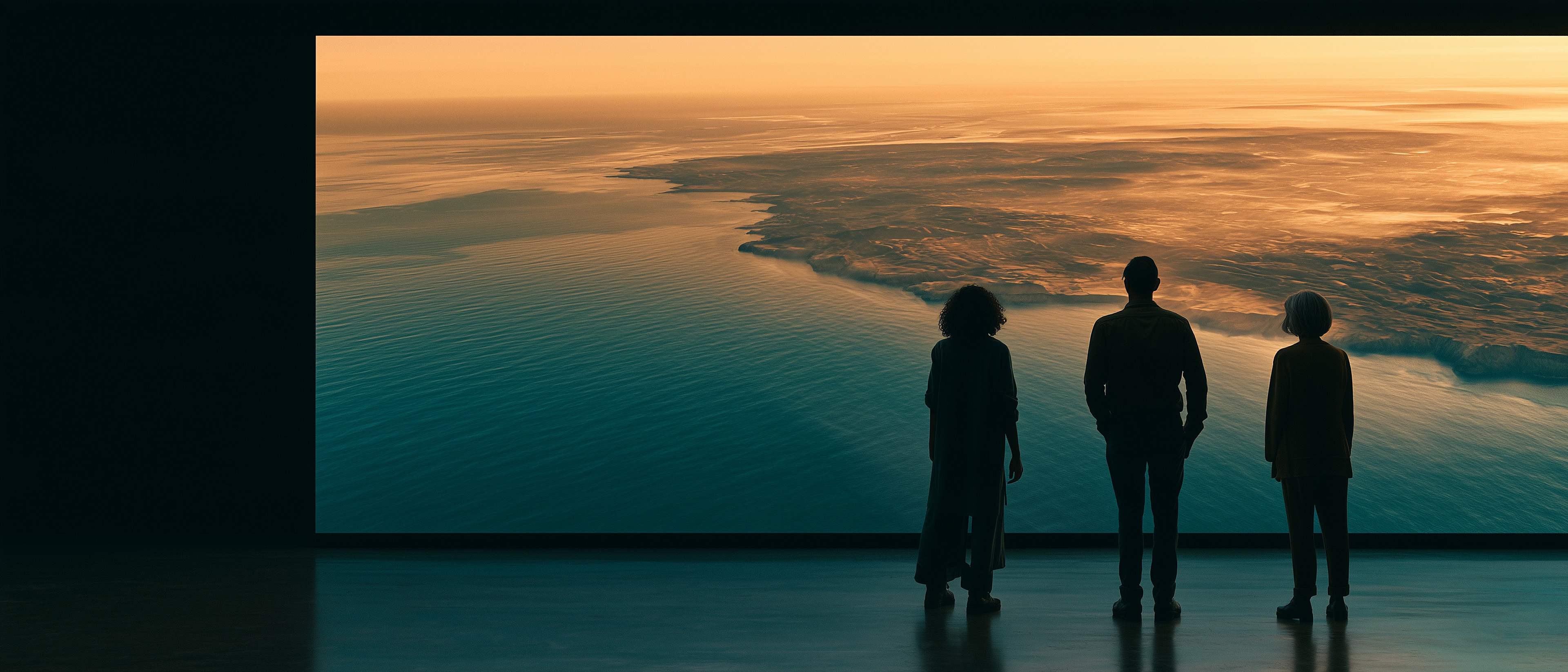 Three colleagues stand in silhouette before an enormous screen showing a coastline at sunrise.